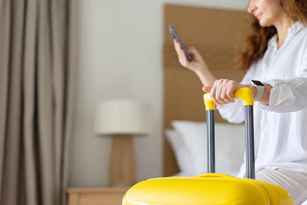 Closeup of adult woman holding suitcase handle while using smartphone in bedroom